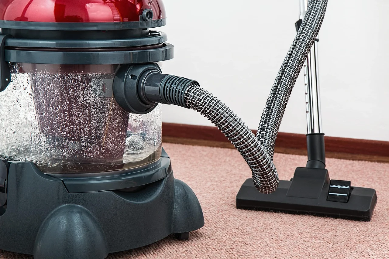 Red and grey canister vacuum cleaner with a clear water filtration drum, corrugated hose and a metal wand with a wide floor brush resting on beige carpet