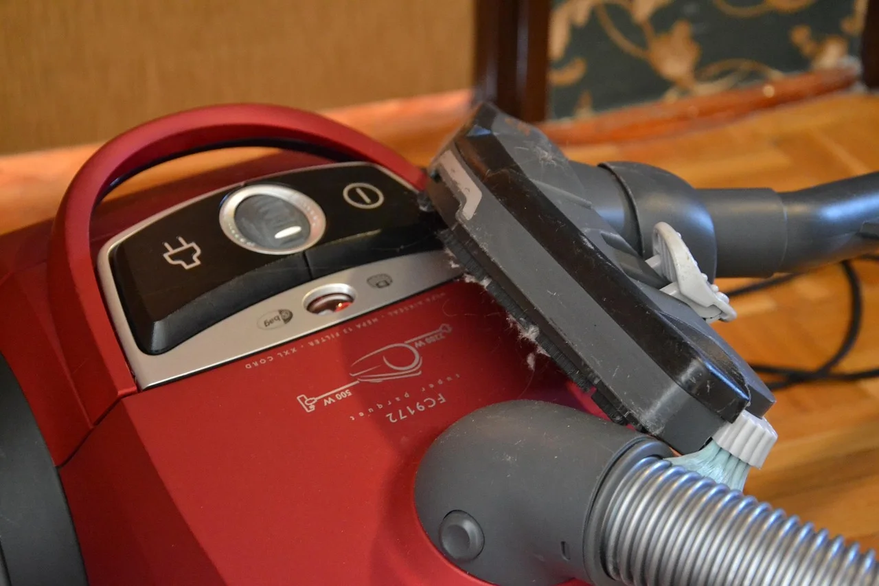 Close-up of a red canister vacuum cleaner's control panel and cord rewind buttons with a dust-clogged floor brush and hose resting on top of it