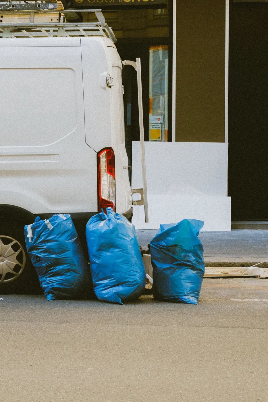 Three full blue waste sacks propped against the rear of a white van at the kerbside