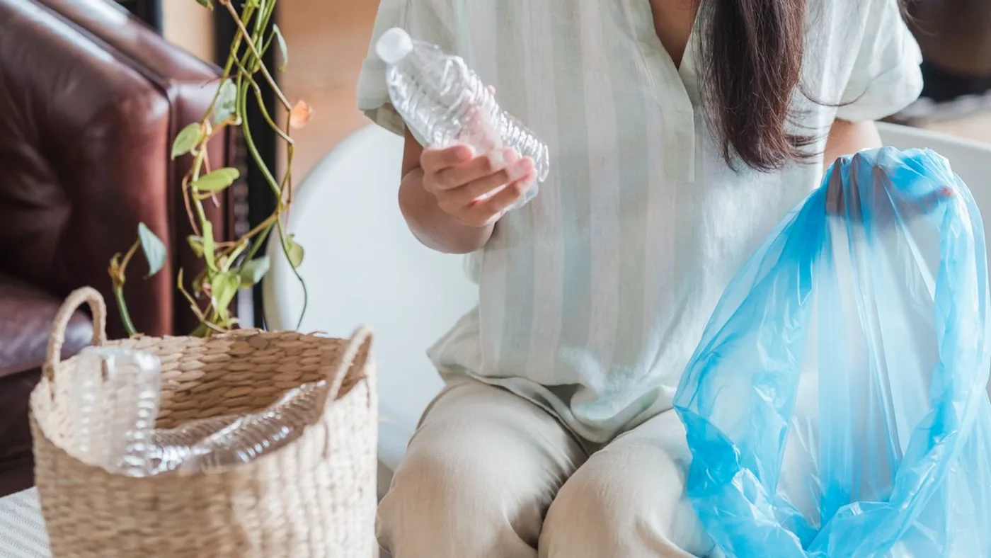 Person sitting at home holding an empty crushed plastic bottle over an open blue bag, beside a woven basket already filled with clear bottles