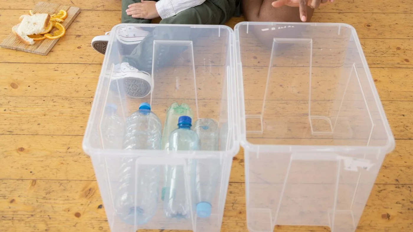 Two clear plastic storage boxes side by side on a wooden kitchen floor, one holding empty drinks bottles and the other left empty