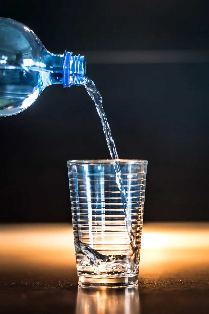 Clear water pouring from a plastic bottle into a cut-glass tumbler on a dark surface with warm light glowing behind the glass