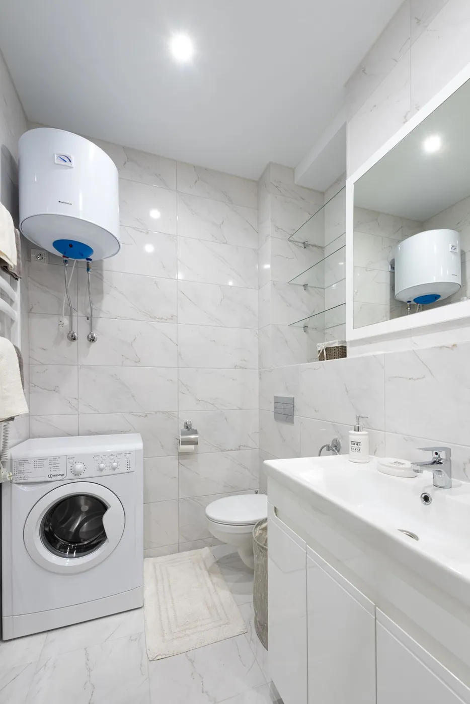 Compact wall-mounted white water heater installed above a washing machine in a tiled bathroom utility corner
