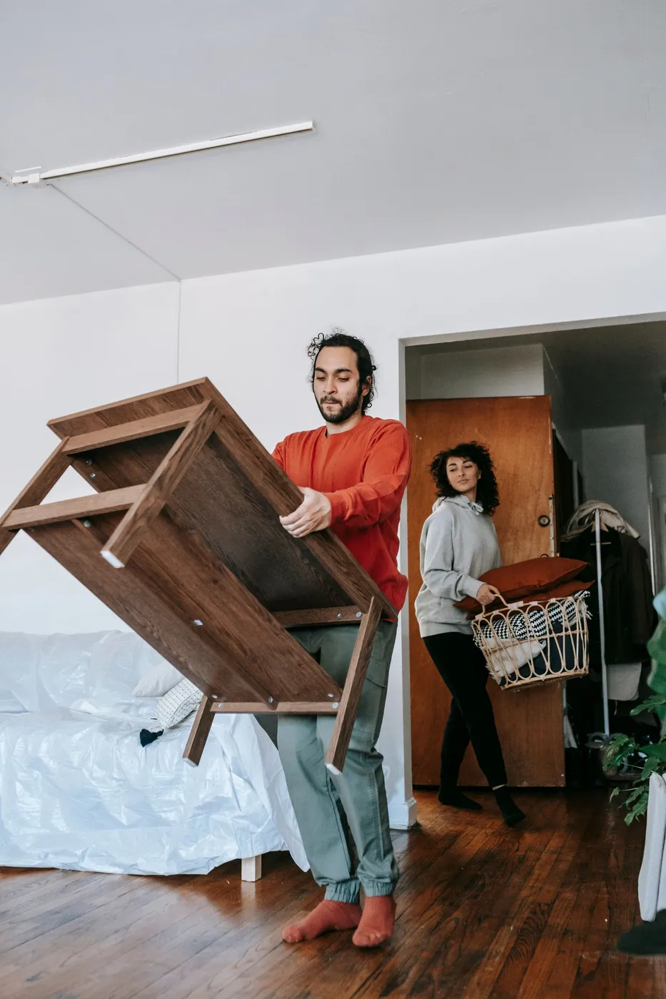 Man in a red top carrying a wooden console table tipped on its side through an apartment while a woman carries a basket of cushions behind him