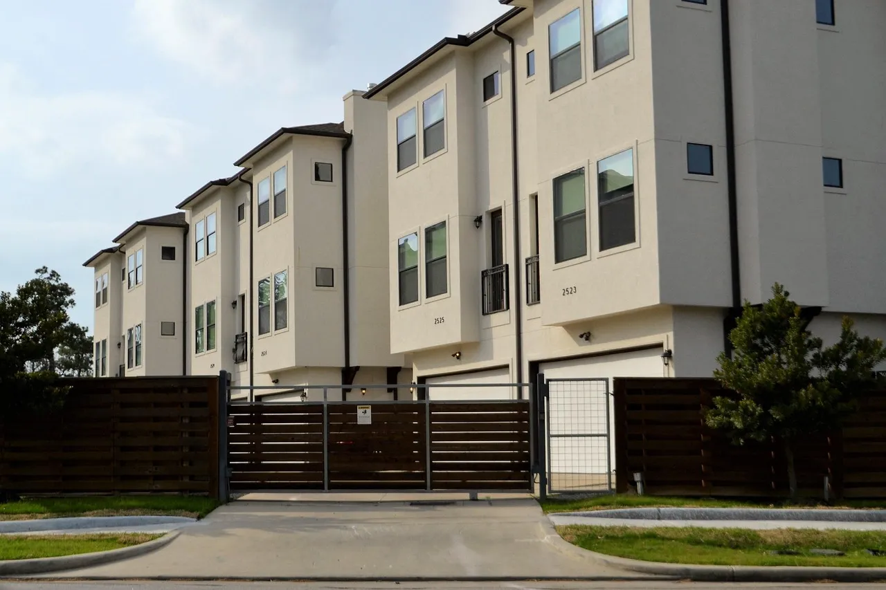 Row of three-storey cream townhouses with private garages behind a slatted wooden community gate and driveway