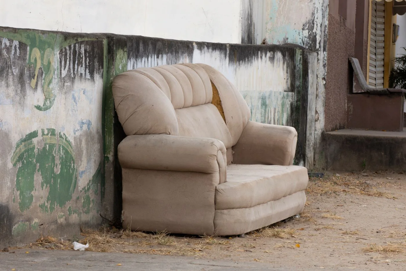Worn beige two-seater sofa with a torn back cushion abandoned against a stained concrete wall on a dusty street