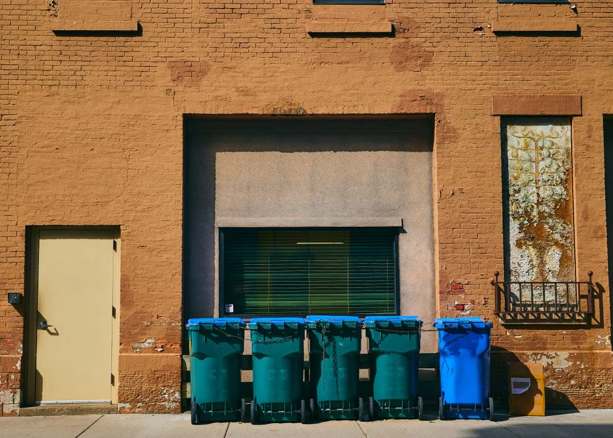 A neat row of residential wheelie bins with blue and green lids lined up outside a building along a sunny sidewalk