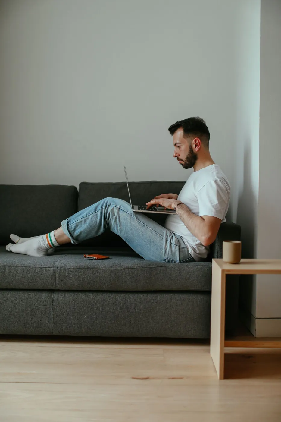 Man sitting on a grey sofa in a minimalist living room, typing on a laptop resting on his lap